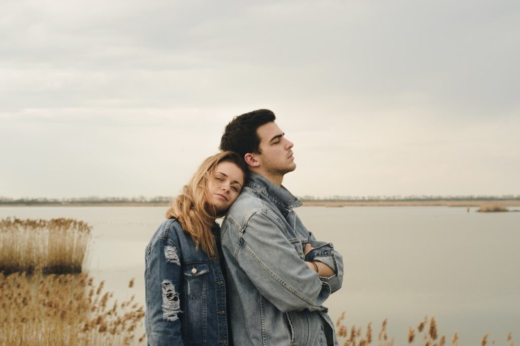 woman in blue denim jacket leaning on man's shoulder near body of water